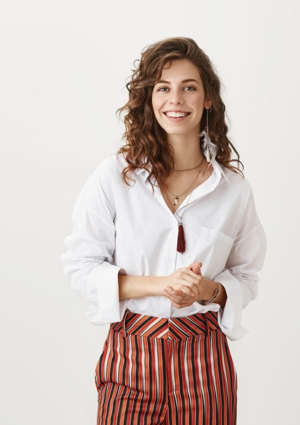 She guarantees good service. Attractive slender woman in trendy striped pants standing over gray background with cheerful smile and holding hands together over chest, trying to control herself.